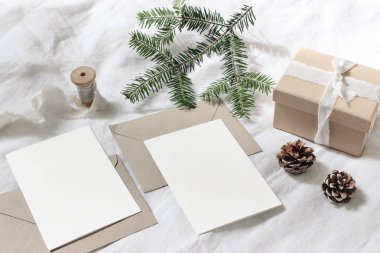 Noel masası, şenlikli kompozisyon. Noel tebrik kartları, zarflar maketleri. Beyaz keten arka plan üzerinde ipek kurdele, çam kozalağı ve köknar ağacı dalı ile el yapımı hediye kutusu. Üstte görüntü.