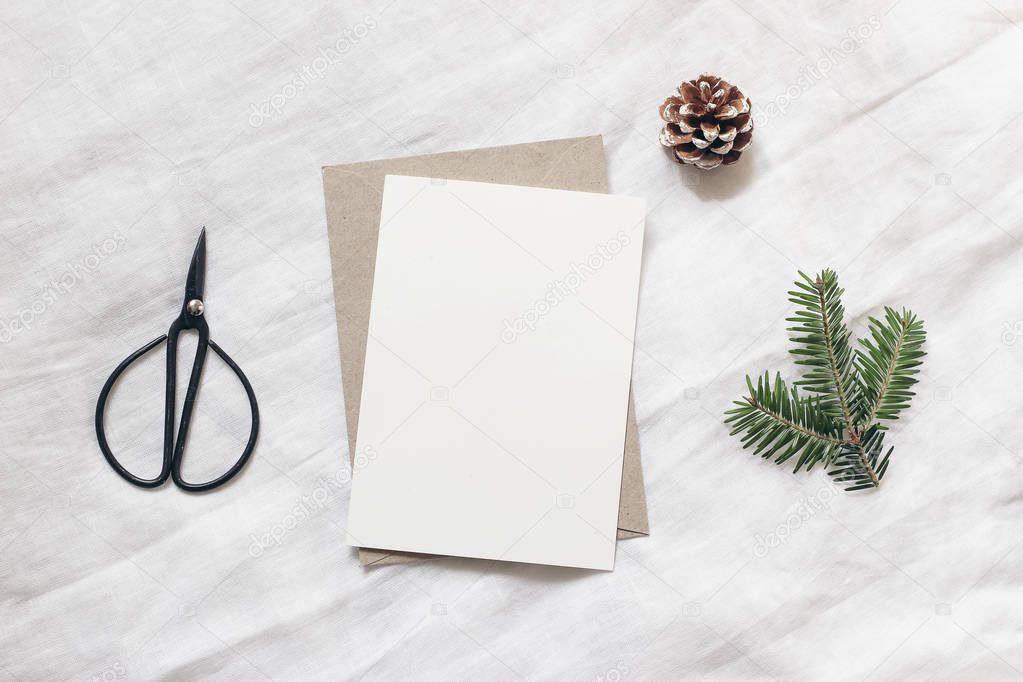 Christmas blank greeting card mock-up scene. Festive winter wedding composition. Envelope, pine cone, black vintage scissors and fir tree branch on white table, linen background. Flat lay, top view.