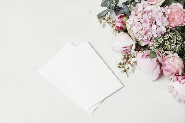 Mariage féminin, scène de maquette d'anniversaire. Coin floral décoratif de pivoine, hortensia, roses et fleurs de sauterelles. Cartes de vœux en papier blanc sur fond de table en bois blanc. Grosse couche, vue du dessus — Photo