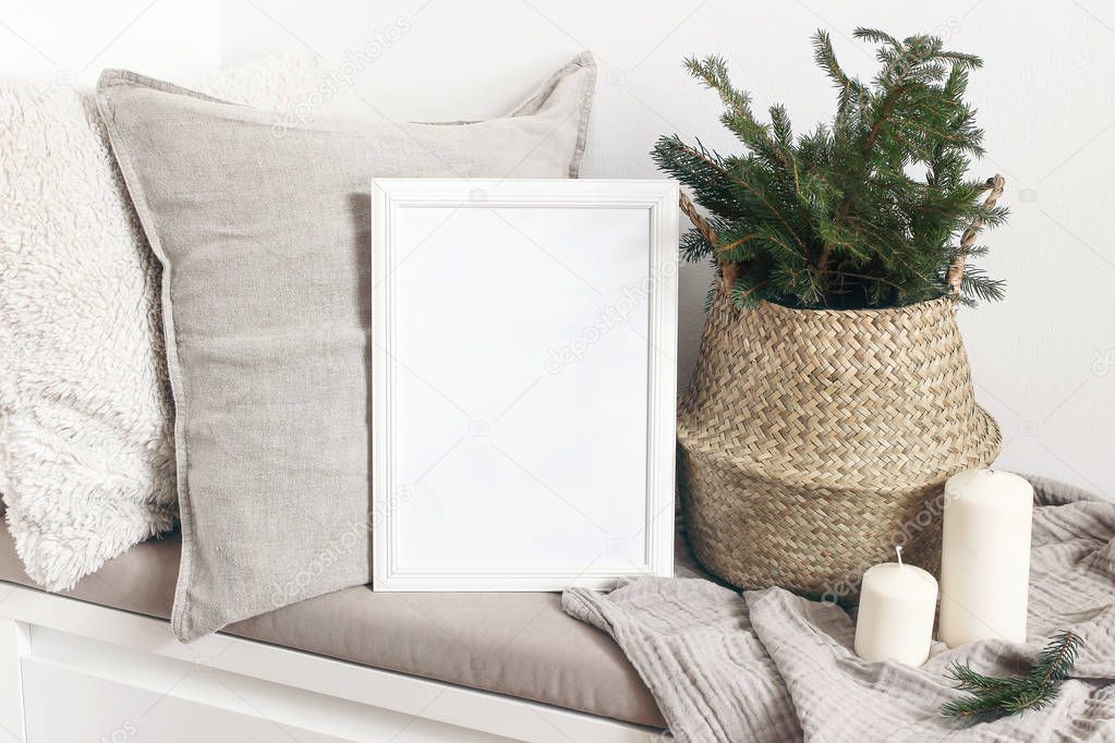White blank wooden frame mockup with Christmas tree, candles, linen cushions and plaid on the white bench. Poster product design. Scandinavian home decor, nordic design. Winter festive concept.