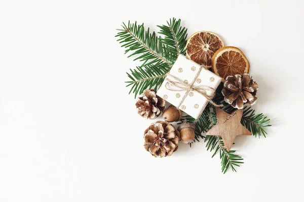 Julstämning stil aktiesammansättning. Handlindade presentförpackning, torra apelsin frukt skivor, ekollon, kottar tall och gran grenar isolerad på vitt bord bakgrund. Vinter platt låg, ovanifrån. — Stockfoto