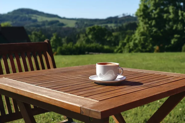 Cup of coffe on wooden table with chair. Blurred green trees and hill tops background. Coffee break in countryside. Picnic, garden party snack, relaxation concept. No people, selective focus.