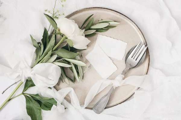 Festive summer wedding scene. Marble table setting with cutlery, olive branches, white peony flowers, stoneware plate and silk ribbon. Blank place cards mockups. Restaurant menu concept. Flat lay, top