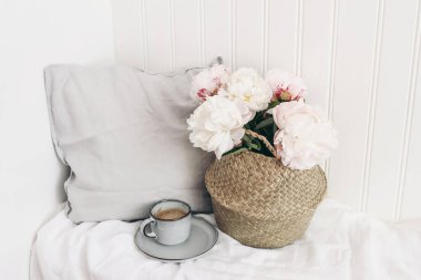 Kadın düğünü ya da doğum günü hayat sahnesi. Saman sepetinde bir buket pembe şakayık, bir fincan kahve, keten yastık. Beyaz ahşap duvar arka planı. Rustik İskandinav iç tasarımı, seçici odaklanma
