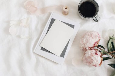 Düğün eşyaları, natürmort. Tebrik kartı, davetiye sahnesi. Bir buket pembe şakayık çiçeği, bir fincan kahve ve beyaz keten masa örtüsü üzerine kitap. Kadınsı fotoğraf, düz yatak, üst görünüm.