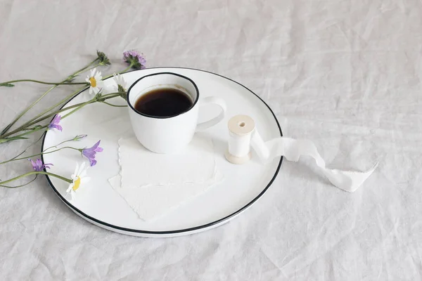 Spring, summer still life scene. Blank cotton paper greeting cards mockups on ceramic plate. Cup of coffee, silk ribbon, daisies and bluebells flowers on linen table cloth. Floral composition, top — Stock Photo, Image