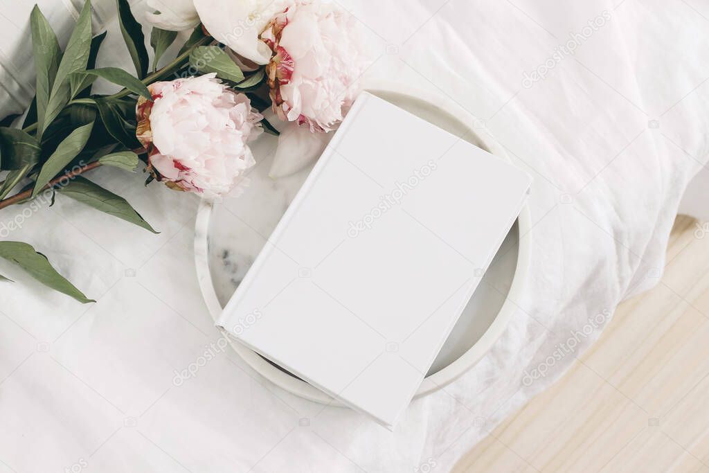 Wedding still life scene. White empty book cover mockup on marble tray. Pink peony flowers on white linen table cloth. Vintage feminine styled photo, wooden floor. Flat lay. Blurred background.