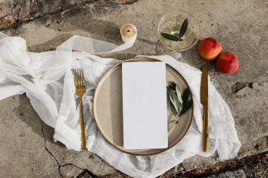 Düğün için yaz sofrası kuracağız. Altın çatal bıçak, zeytin dalı, bir bardak şarap, şeftali meyvesi ve grunge beton arka planda porselen yemek tabağı. Boş menü kartı modeli. Düz yatış, üst görünüm.