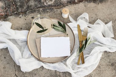 Festive wedding summer table setting. Golden cutlery, olive branch, glass of wine and envelope on porcelain dinner plate. Grunge concrete background. Blank card, invittation mockup. Flat lay, top view clipart