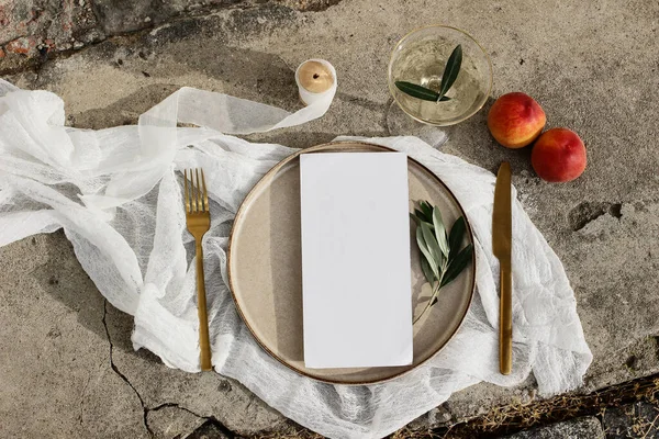 Festive wedding summer table setting. Golden cutlery, olive branch, glass of wine, peaches fruit and porcelain dinner plate on grunge concrete background. Blank menu card mockup. Flat lay, top view. — Stock Photo, Image