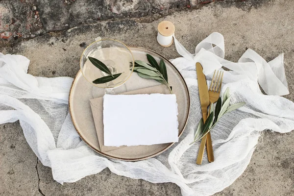 Festive wedding summer table setting. Golden cutlery, olive branch, glass of wine and envelope on porcelain dinner plate. Grunge concrete background. Blank card, invittation mockup. Flat lay, top view — Stock Photo, Image