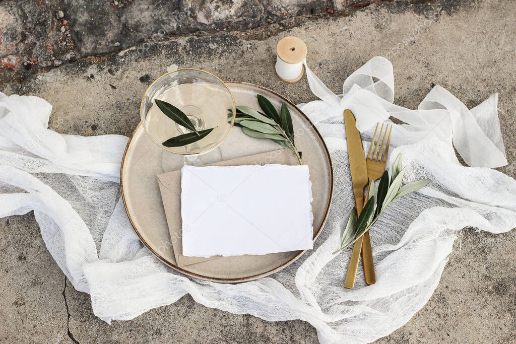 Festive wedding summer table setting. Golden cutlery, olive branch, glass of wine and envelope on porcelain dinner plate. Grunge concrete background. Blank card, invittation mockup. Flat lay, top view