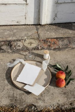 Summer wedding still life scene. Blank card, invitation mock-up. Olive branches, silk ribbon and peaches fruit on grunge concrete background. Blurred old white door. Wine champagne glass, vertical clipart