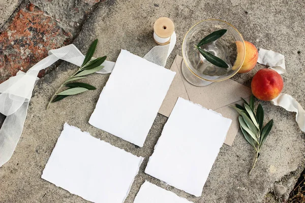 Summer wedding stationery, still life scene. Blank cards, invitations mock-ups set. Olive branches, silk ribbon, peaches fruit and champagne wine drink, glass on grunge concrete background. Flat lay