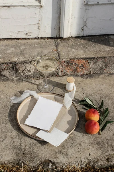 Summer wedding still life scene. Blank card, invitation mock-up. Olive branches, silk ribbon and peaches fruit on grunge concrete background. Blurred old white door. Wine champagne glass, vertical — Stock Photo, Image