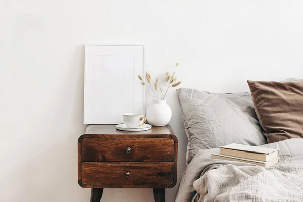 Portrait white frame mockup on retro wooden bedside table. Modern white ceramic vase with dry Lagurus ovatus grass and cup of coffee. Beige linen and velvet pillows in bedroom. Scandinavian interior. — Stock Photo, Image
