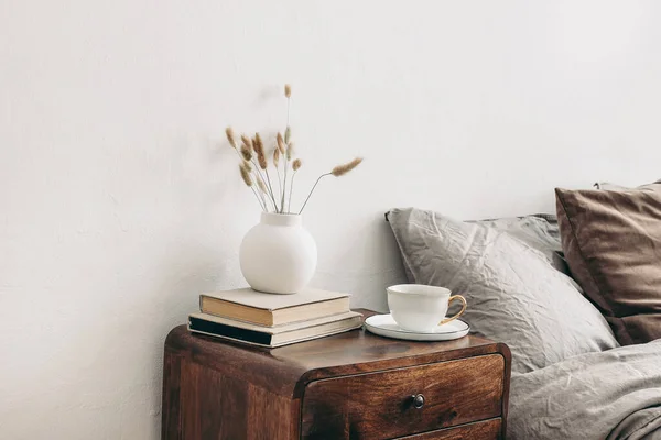 Moderní bílá keramická váza se suchou trávou Lagurus ovatus a šálkem kávy na retro dřevěném nočním stolku. Béžové prádlo a sametové polštáře v ložnici. Skandinávský interiér. Homestaging. — Stock fotografie