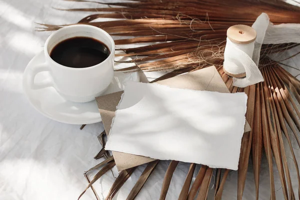 Sommer Hochzeit Papeterie Attrappe-Szene. Leere Grußkarte, Umschläge, Seidenband und trockenes Palmblatt im Sonnenlicht. Tasse Kaffee auf weißem Tischhintergrund mit Schatten. Hoher Winkel, Draufsicht. — Stockfoto