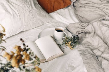 Sonbahar kahvaltısı hala hayattadır. Bir fincan kahve, açık bir kitap ve beyaz keten arka planda tansi çiçekler. Bulanık çiçekli ön plan kadife ve yatakta keten yastıklar. Huysuz sonbahar yaşam tarzı konsepti. Üst görünüm.