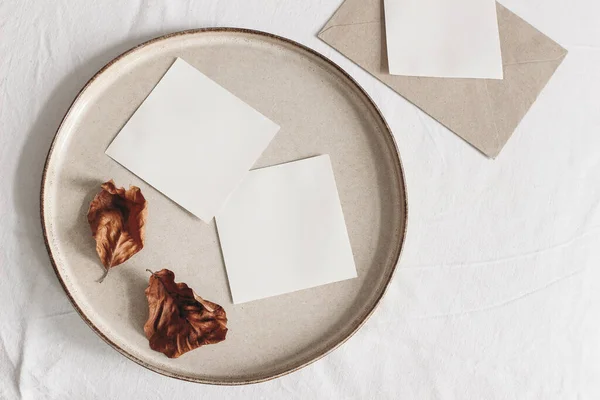 Autumn composition. Blank memo cards mockups, craft envelope and dry beech leaves on white linen table cloth background. Fall and Thanksgiving concept. Styled flat lay photography. Top view.