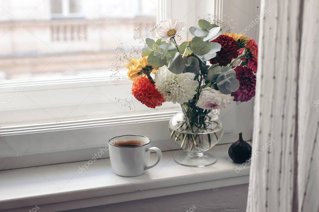 Feminine autumn still life. Cup of coffee, bouquet of colorful dahlia flowers in glass vase on windowsill. Styled moody floral composition with fig fruit. Window with white curtain. Selective focus.
