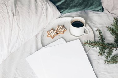 Noel şenliği natürmort. Boş kağıtlar. Bir fincan kahve, zencefilli kurabiye ve seramik tabakta ladin ağacı dallarıyla davetiye maketleri. Tatil geçmişi. Yatakta kahvaltı. Üst görünüm.