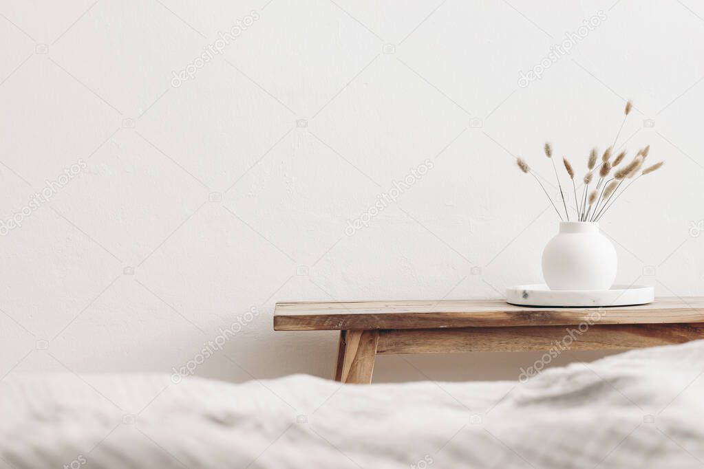 Modern white ceramic vase with dry Lagurus ovatus grass and marble tray on vintage wooden bench, table. Blurred beige linen blanket in front. Scandinavian interior. Empty white wall, copy space.