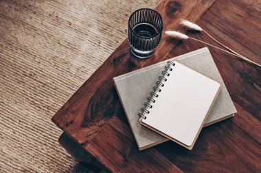 Boş günlük, not defteri modeli ve ahşap masaya kitap. Bir bardak su ve kuru Lagurus ovatus otu. Bulanık bej halı arka planı. Düz yerde, üst tarafta. Basit İskandinav boho iç mimarisi.
