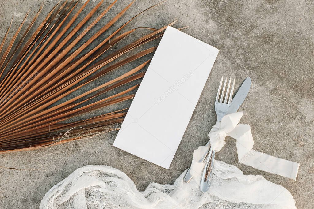 Tropical wedding ry still life. Closeup of blank menu card mock-up. Dry palm leaf, silver cutlery and muslin runner on grunge beige concrete background. Moody boho design. Flat lay, top view.