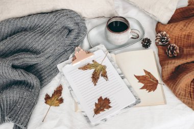 Sonbahar kahvaltısı hala hayattadır. Bir fincan kahve, açık kitap ve renkli akçaağaç ve meşe yapraklı not defteri. Beyaz keten arka plan. Yatakta keten yastıklar ve gri süveter. Sonbahar yaşam tarzı konsepti. Üst görünüm.