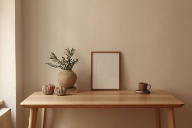 Autumn interior still life. Vase with green olive tree branches, pumpkins on table. Cup of tea, coffee, books. Wooden poster, picture frame mockup. Home office. Scandi design, beige wall background.