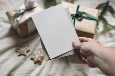 Christmas still life. Female hand holding blank vertical greeting card, invitation mockup. Blurred gift boxes, gingerbread cookies, fir tree branches. White linen tablecloth. Winter composition, top