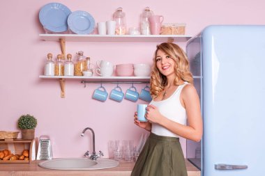 Genç mutlu kadın mutfakta evde kahve veya çay içiyor. Ofis gitmeden önce onu kahvaltı sahip sarışın güzel kız. Kahve molası. Pembe ve mavi pastel modern mutfak iç