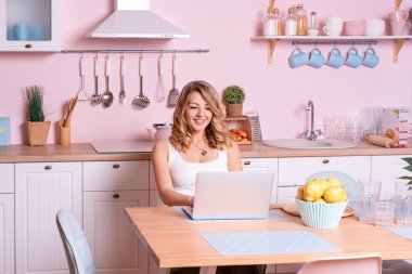 Evde mutfakta laptop kullanarak gülümseyen genç kadın. Sarışın kadın bilgisayar üzerinde çalışır, Freelancer veya Blogger evde çalışan