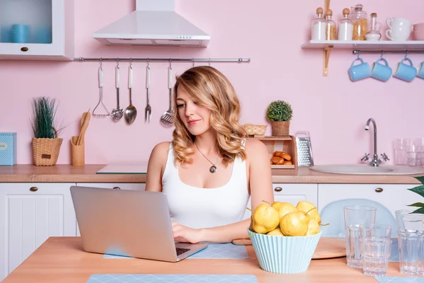 Lächelnde junge Frau mit Laptop in der Küche zu Hause. Blondine arbeitet am Computer, Freelancer oder Bloggerin zu Hause — Stockfoto