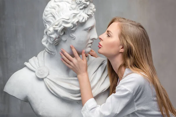 Giovane donna baciare Dio Apollo busto scultura. Antico dio greco del sole e della poesia Copia in gesso di una statua di marmo su sfondo murale in cemento grandioso. Arte antica e bellezza vivente — Foto Stock