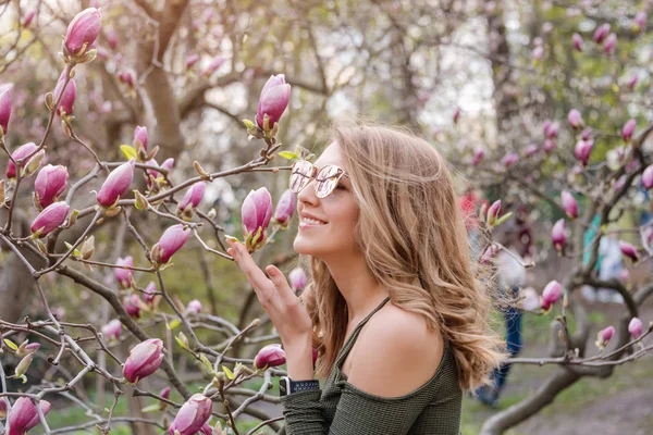 Молодая блондинка рядом с цветущим цветочным деревом магнолии в весеннем парке в солнечный день. Деревья Магнолии. Красивая счастливая девушка наслаждается запахом в цветущем весеннем саду — стоковое фото