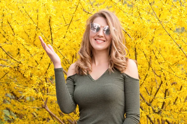 Giovane donna bionda vicino agli alberi gialli in fiore nel parco primaverile nella giornata di sole. Bella ragazza felice godendo odore in un giardino fiorito primavera — Foto Stock