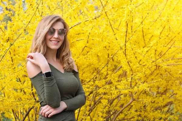 Giovane donna bionda vicino agli alberi gialli in fiore nel parco primaverile nella giornata di sole. Bella ragazza felice godendo odore in un giardino fiorito primavera — Foto Stock