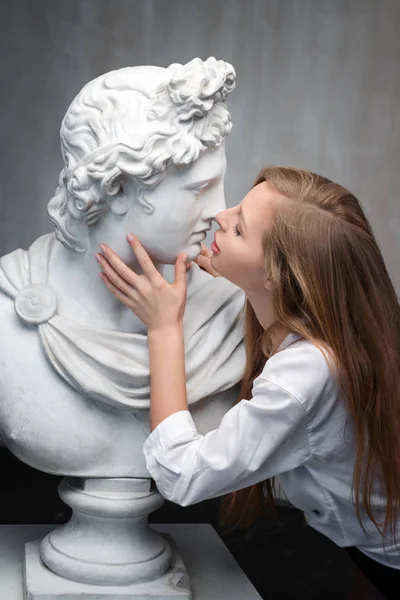 Giovane donna baciare Dio Apollo busto scultura. Antico dio greco del sole e della poesia Copia in gesso di una statua di marmo su sfondo murale in cemento grandioso. Arte antica e bellezza vivente — Foto Stock