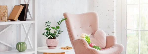Stylish pink armchair with heart shaped pillow in a bright minimalist interior. Wooden shelving unit with decor near grey wall. Bookcase with candles, living room interior details