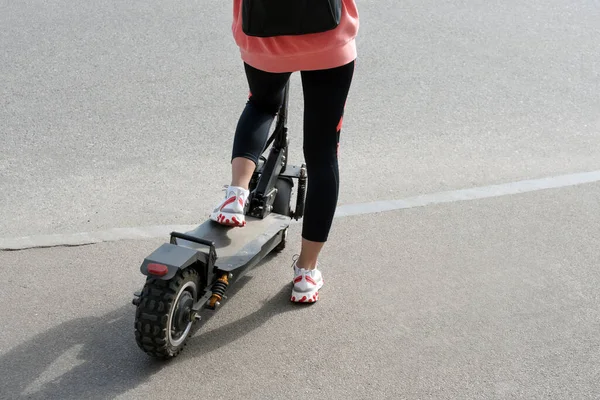 The legs of an unknown girl in white sneakers and gym leggings riding on a black electric scooter over urban asphalt. Modern transportation, electric kick scooter
