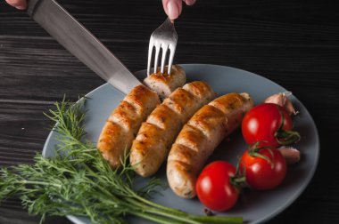 Üç ızgara sosis ahşap siyah arka plan üzerinde gri bir tabakta da kiraz domates, yeşillik, sarımsak, bıçak, çatal yalan. Yukarıdan görüntüleyin. Metin ve logo için yer.