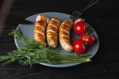 Üç ızgara sosis ahşap siyah arka plan üzerinde gri bir tabakta da kiraz domates, yeşillik, sarımsak, bıçak, çatal yalan. Yukarıdan görüntüleyin. Metin ve logo için yer.