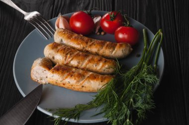 Üç ızgara sosis ahşap siyah arka plan üzerinde gri bir tabakta da kiraz domates, yeşillik, sarımsak, bıçak, çatal yalan. Yukarıdan görüntüleyin. Metin ve logo için yer.