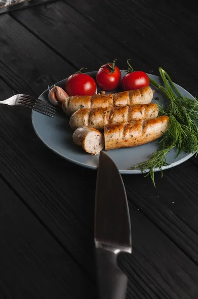 Drie gegrilde worstjes op een grijze plaat op een houten zwarte achtergrond liggen ook een cherry tomaat, Groenen, knoflook, mes, vork. Bekijk van bovenaf. Plek voor tekst en logo. — Stockfoto