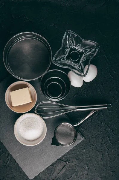 Ingredients and dishes on a black concrete table, where the corolla also lies, is filled with flour, three chicken eggs and a slate board. View from above. Place under the text