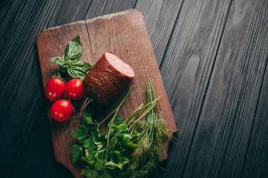 Siyah bir tablo ve bir kahverengi tahta üzerinde füme salam. Biberiye, sarımsak ve biber ve domates kiraz, dereotu, maydanoz ile sosis