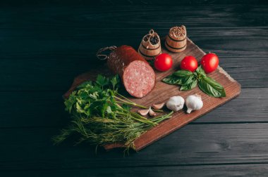 Siyah bir tablo ve bir kahverengi tahta üzerinde füme salam. Biberiye, sarımsak ve biber ve domates kiraz, dereotu, maydanoz ile sosis