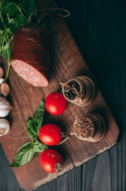 Siyah bir tablo ve bir kahverengi tahta üzerinde füme salam. Biberiye, sarımsak ve biber ve domates kiraz, dereotu, maydanoz ile sosis